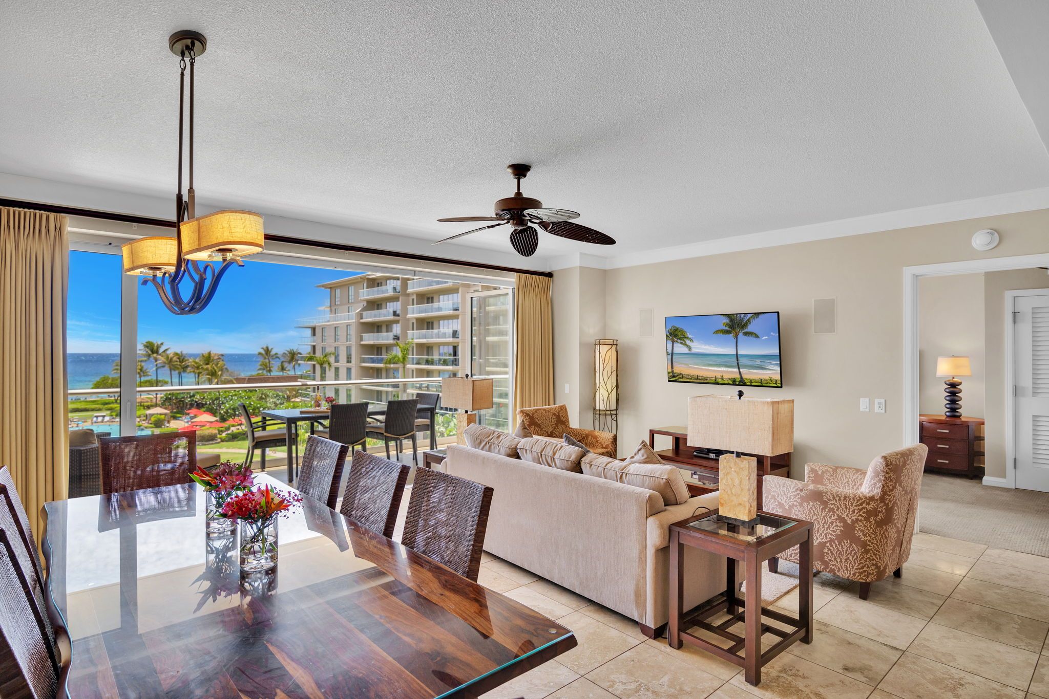 A modern hotel room with a dining area, a sofa set, a ceiling fan, and a large window with a view of the ocean.