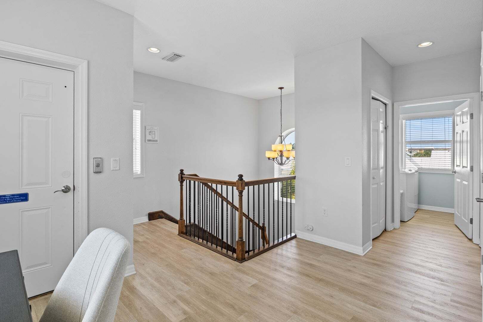 A hallway with wooden flooring, white walls, a staircase with a wooden railing, and a chandelier.