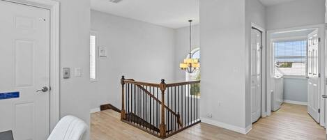 A hallway with wooden flooring, white walls, a staircase with a wooden railing, and a chandelier.