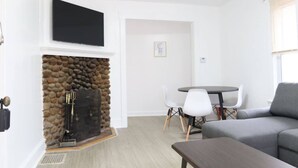 A living room with a stone fireplace, a flat-screen TV, a grey sofa, a round table with chairs, and a window with blinds.