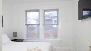 A bedroom with a bed, two windows, a nightstand, a lamp, and a TV mounted on the wall.