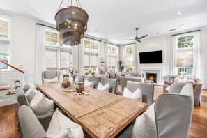 A spacious living room with a large wooden dining table, comfortable seating, a fireplace, and a television mounted on the wall. - 5BR WaterColor Retreat:  Beach Club Access, Golf Cart, BIkes, Private Pool, Carriage House! (Santa Rosa Beach)
