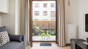 En skjutdörr som leder ut till en balkong med ett bord och stolar, en grå soffa, en liten pall och ett element.