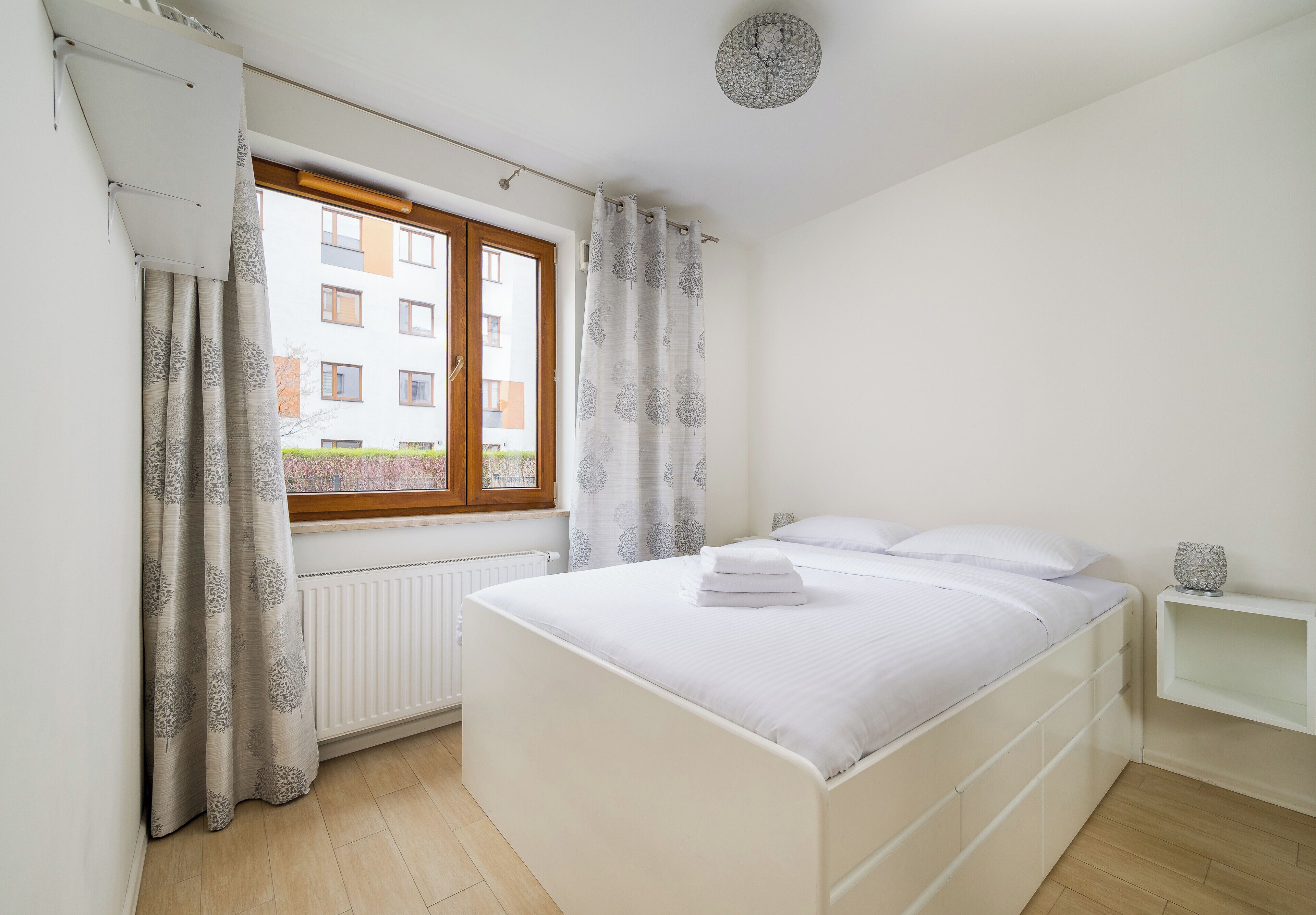 Quarto com cama, janela, radiador e prateleira fixada na parede.