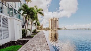 A waterfront walkway with a pool, palm trees, and modern buildings. - Luxury Waterfront Studio Condo with Balcony Views (Fort Myers)
