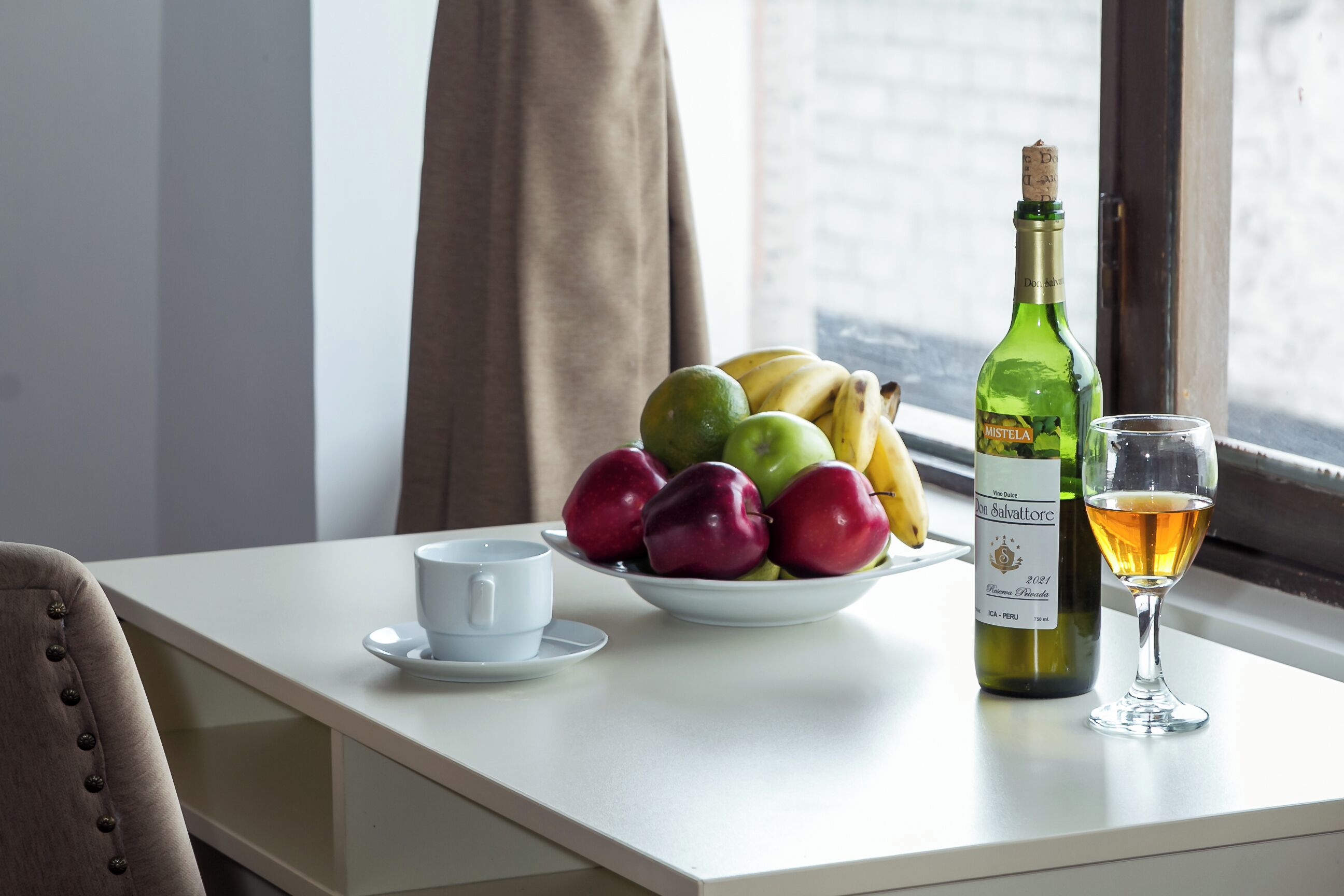 A table with a bowl of fruit, a wine bottle, a wine glass, and a cup.