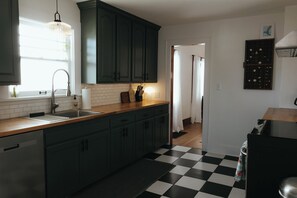 Una cocina con armarios verde oscuro, encimera de madera, suelo a cuadros blanco y negro, lavavajillas de acero inoxidable y una lámpara colgante.