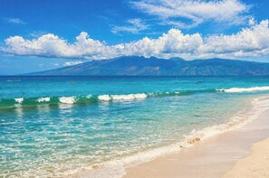 Ein Strand mit klarem türkisblauem Wasser, einem Sandufer und einer Bergkette im Hintergrund.