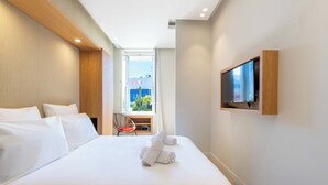 A hotel room with a large bed, a TV mounted on the wall, a chair, and a view of the cityscape through the window. - 2-Br Facing Palais des Congrès – Cannes City Centre (Cannes)