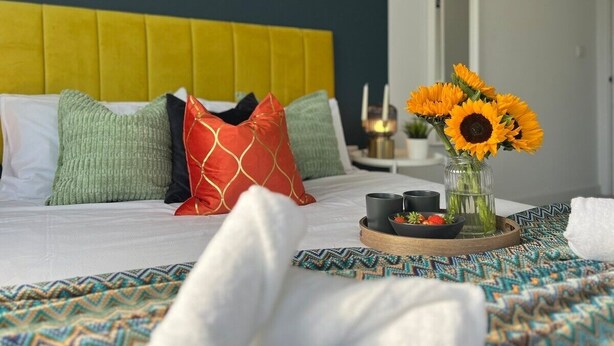 Ein ordentlich bezogenes Bett mit Kissen, ein Tablett mit Speisen und Getränken sowie eine Vase mit Sonnenblumen.