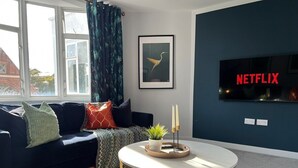 A living room with a sofa, a round coffee table, a framed picture, a television, and a window with curtains. - Welcome to The Aviary – Upper (Bournemouth)