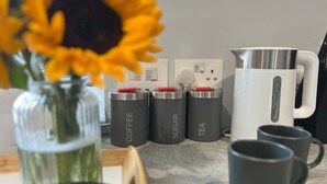 A kitchen counter with labeled containers for coffee, sugar, and tea, a white electric kettle, and two black mugs. - Welcome to The Aviary – Upper (Bournemouth)