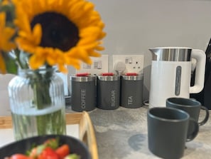 Eine Küchenarbeitsplatte mit beschrifteten Behältern für Kaffee, Zucker und Tee, ein weißer Wasserkocher und zwei schwarze Tassen.