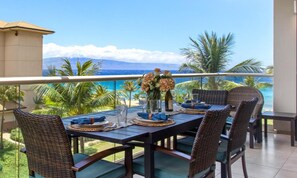 Un coin repas avec une table dressée pour quatre personnes, offrant une vue sur la côte, avec des palmiers et une montagne en arrière-plan.