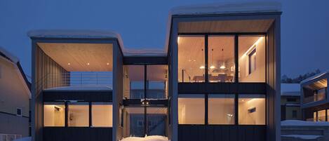 A modern two-story house with large windows and a balcony, surrounded by snow.