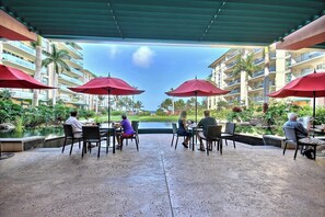 An outdoor dining area with tables and chairs, red umbrellas, and a view of a pool and buildings. - KBM Resorts | 4 FREE CARS | Honua Kai Hokulani | Ocean view | 7-Bedroom Multi-Unit Listing Recently Remodeled! HK ML-2939 (Lahaina)