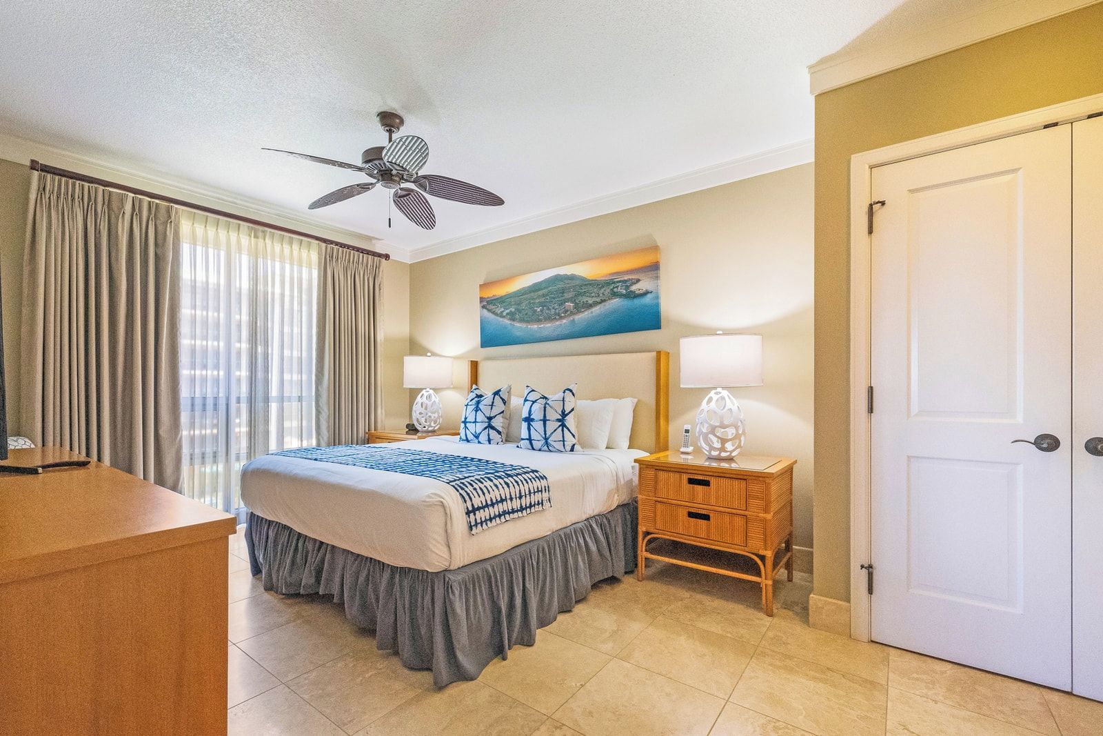 Une chambre d’hôtel avec un lit, un ventilateur de plafond, un tableau, un bureau et une porte.