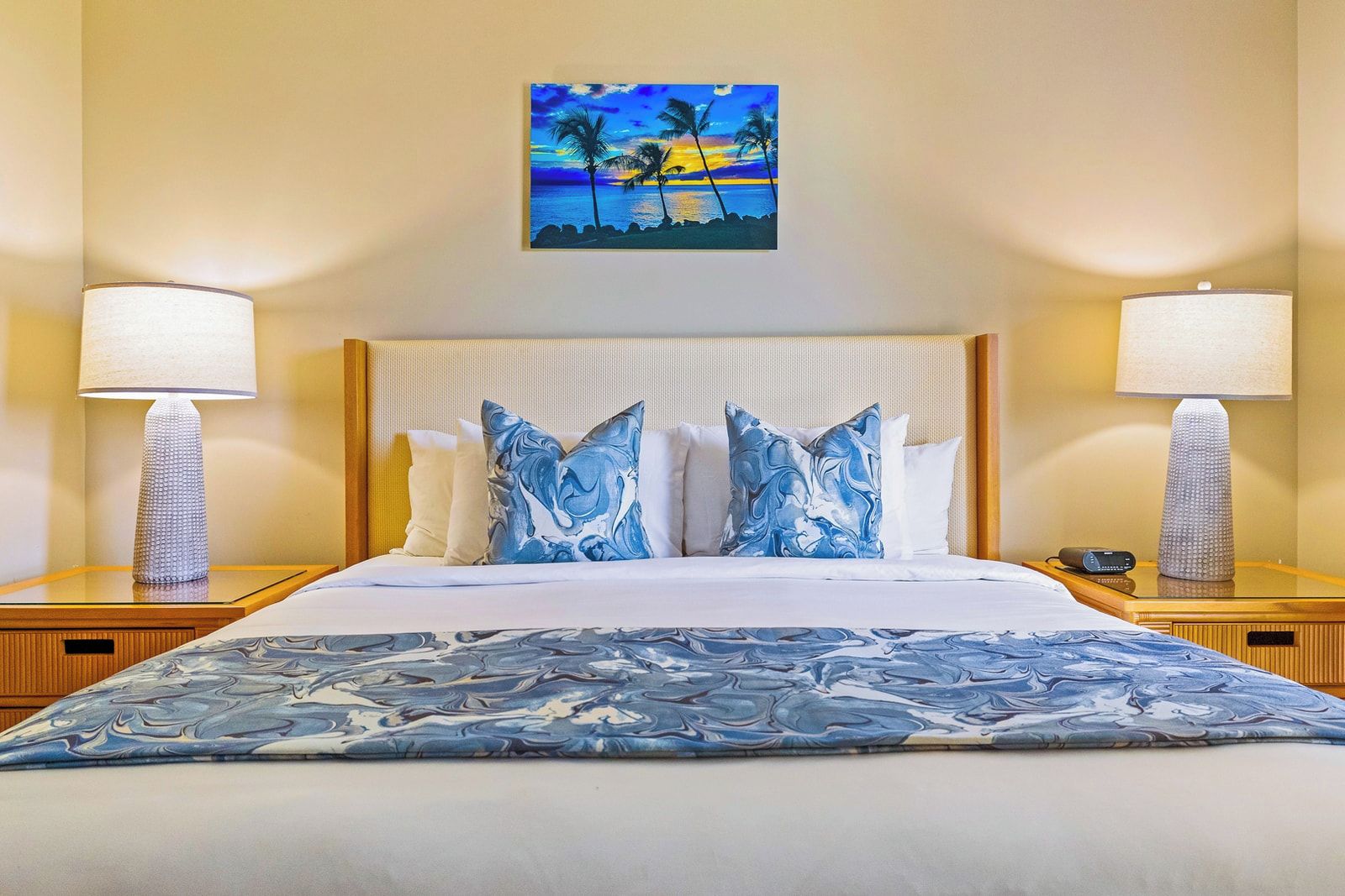 A hotel room with a bed, two bedside lamps, a painting of a sunset, and a patterned bedspread.