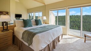 Un dormitorio con una cama grande, televisión, ventilador de techo y vistas a un bosque a través de amplios ventanales.