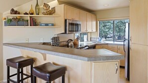 Una cocina con una isla grande, taburetes de bar, armarios de madera, un microondas y un refrigerador.