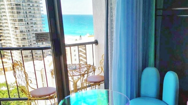 A balcony with a view of the ocean, a glass table, and two chairs. - Blue Bunny-Eared Stay | Ocean View | 2min to Beach (Surfers Paradise)
