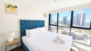 Ein Hotelzimmer mit einem großen Bett, einem Nachttisch, einer Lampe und Blick auf die Stadt durch große Fenster.