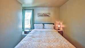 Een slaapkamer met een bed, nachtkastje, lamp, raam met gordijnen en een schilderij van bergen aan de muur.
