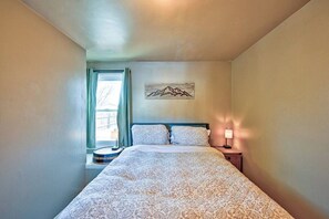 Ein Schlafzimmer mit einem Bett, einem Nachttisch, einer Lampe, einem Fenster mit Vorhängen und einem Wandbild von Bergen.