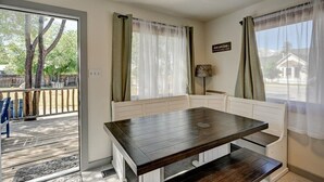 Een kamer met een donker houten tafel en een witte bank, een groot raam met dunne gordijnen, en uitzicht op een terras en bomen.