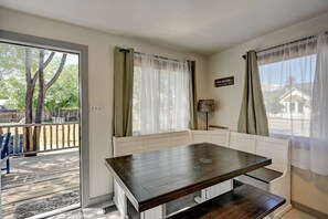 Ein Zimmer mit einem dunklen Holztisch und weißen Sitzbänken, einem großen Fenster mit transparenten Vorhängen und Blick auf eine Terrasse und Bäume.