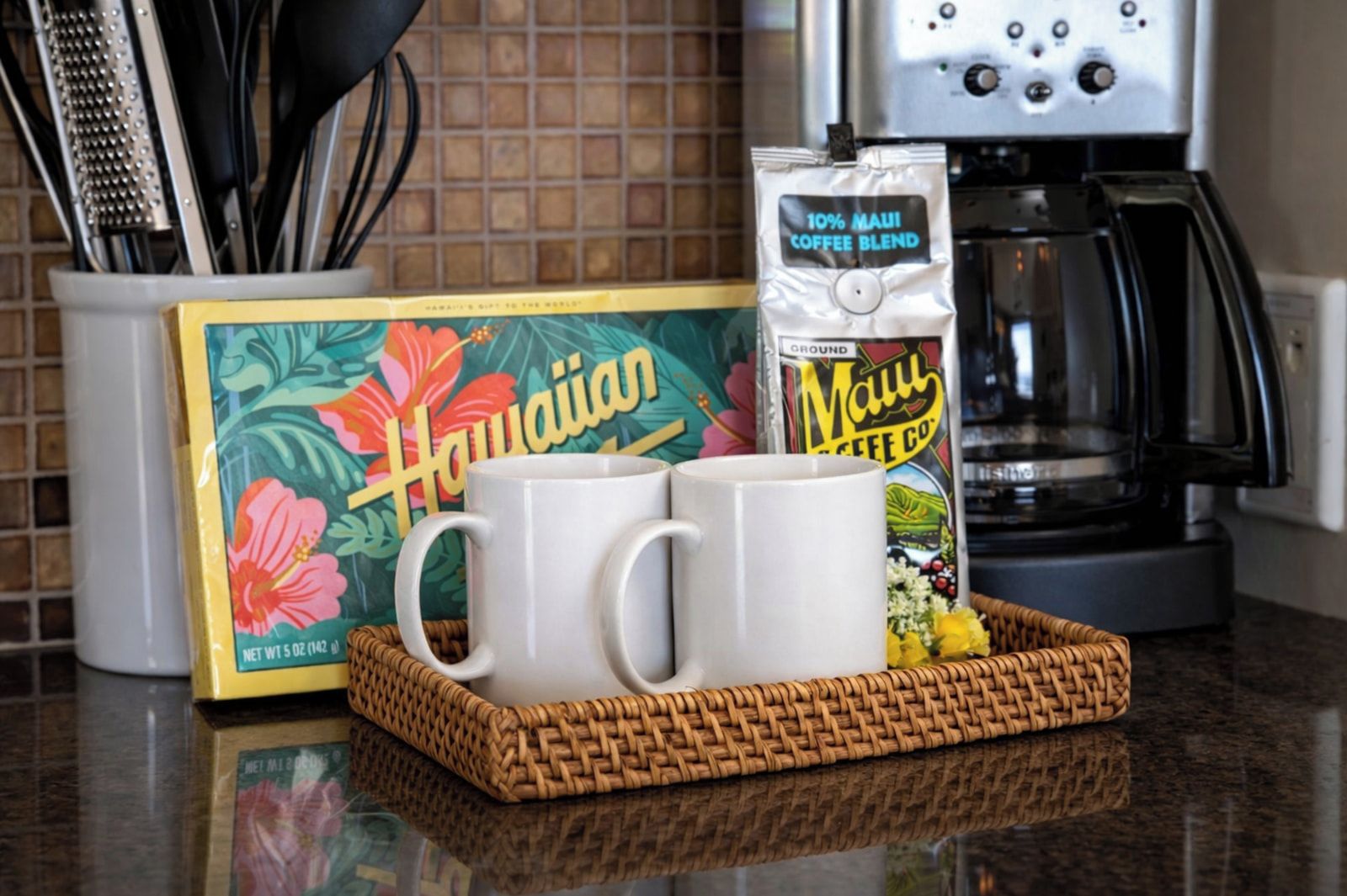Un comptoir de cuisine avec une machine à café, une boîte étiquetée « Hawaï », un sac de café étiqueté « Maui Coffee Co. » et une tasse blanche.