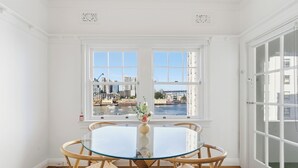 Una habitación con una mesa redonda de vidrio, dos ventanas con vistas a la ciudad y una puerta con paneles de vidrio.