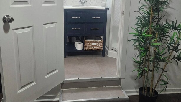 A hallway leading to a bathroom with a dark blue vanity, two wall-mounted lights, a potted plant, and a white door.