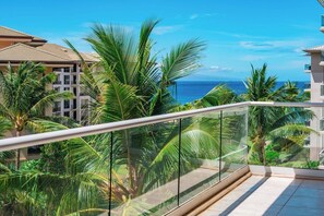 Varanda com grades de vidro com vista para paisagem tropical, palmeiras e o oceano.