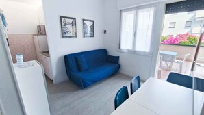 Et lille værelse med tekøkken, en blå sofa, et spisebord med stole og et vindue med gardiner.