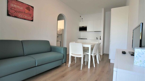 Un salon moderne avec un canapé de couleur turquoise, une table à manger avec des chaises blanches, un coin cuisine avec des meubles blancs et un téléviseur posé sur un meuble blanc.