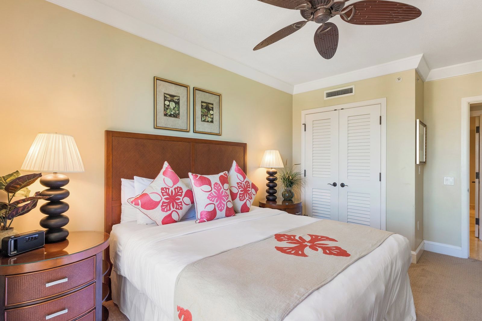 Habitación de hotel con cama, dos lámparas de noche, ventilador de techo, mesita de noche, cómoda y una puerta, además de dos cuadros en la pared.