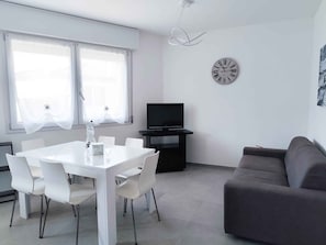 A modern living room with a white dining table, grey sofa, and a flat-screen TV. - Holiday apartment with pool (Cavallino-Treporti)