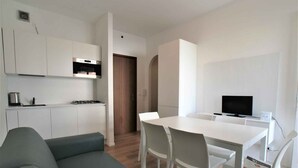 Una cucina moderna con mobili bianchi e piano cottura integrato, una zona pranzo con tavolo e sedie bianche, e un divano grigio.