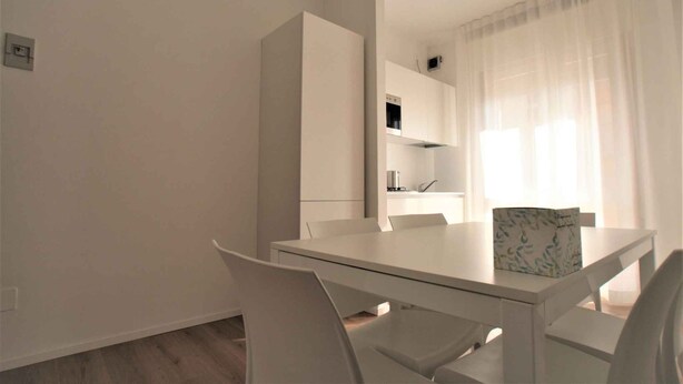 Zona pranzo moderna con tavolo e sedie bianche, una cucina sullo sfondo e tende leggere alla finestra.