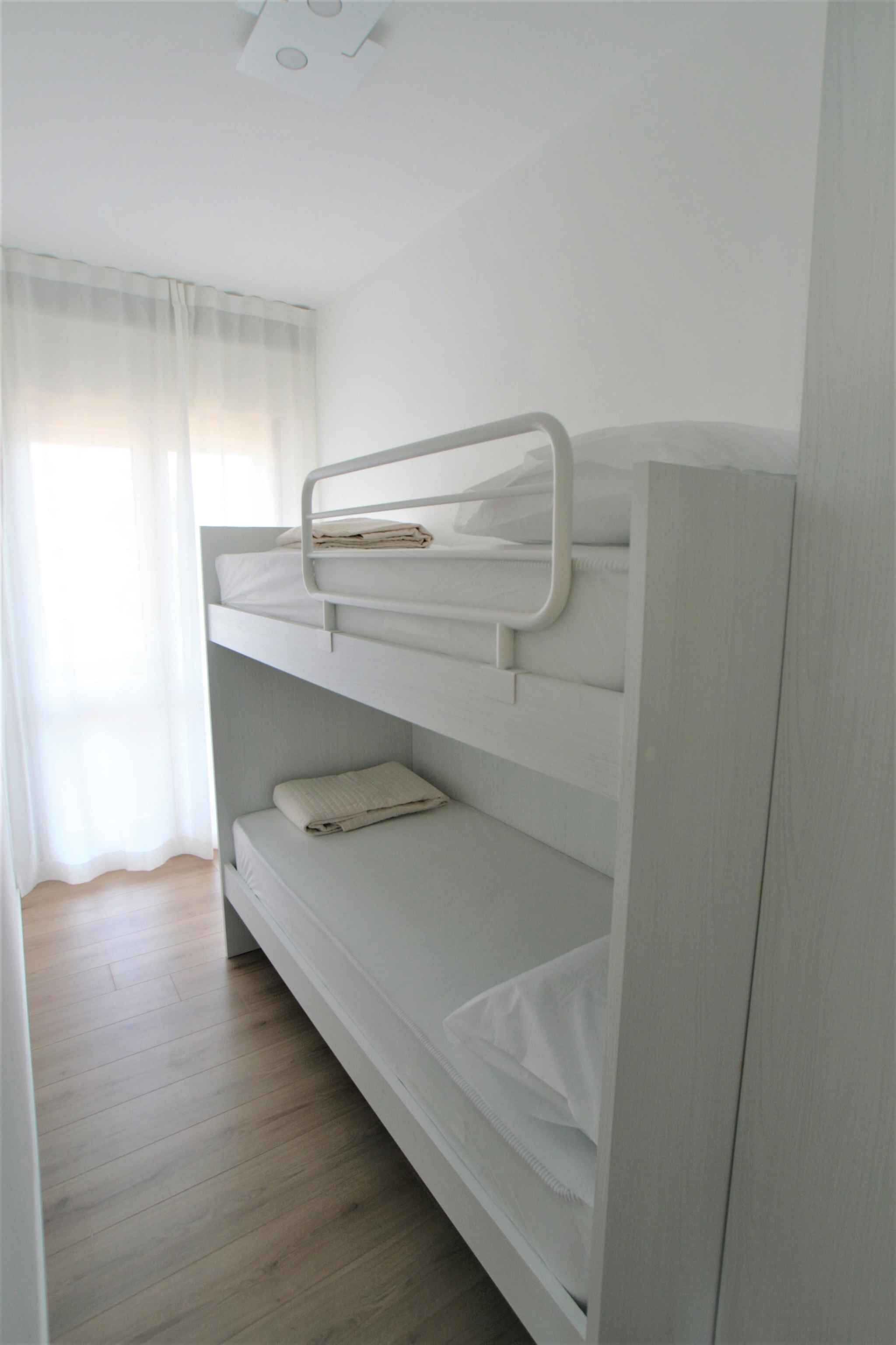 Una cama litera con ropa blanca y estructura de metal, suelo de madera y ventana con cortinas de encaje.