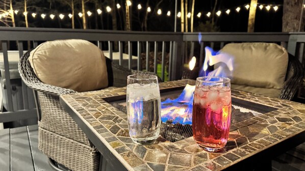 A patio with two glasses of iced beverages, a fire feature, wicker furniture, and string lights.