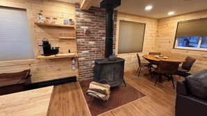 A cozy cabin interior with a wood-burning stove, wooden shelves with coffee equipment, a dining table with chairs, and a window with a view of the outdoors.