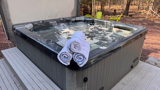 A hot tub with rolled towels on top, surrounded by a wooden deck and a forested area.