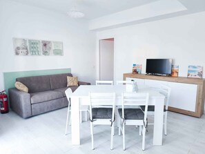 A modern living room with a grey sofa, white dining table, and chairs, a TV on a wooden stand, and a fire extinguisher. - Holiday flat with balcony (Cavallino-Treporti)