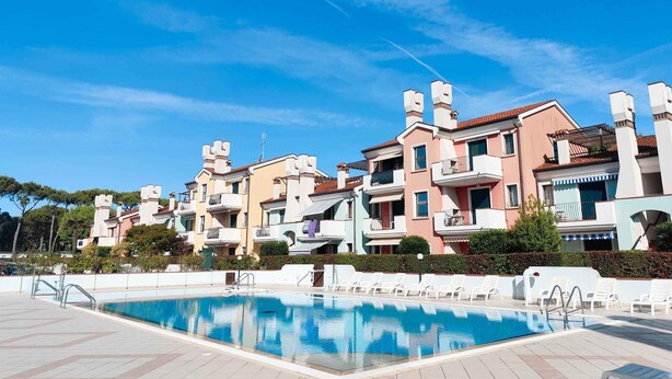 A swimming pool area with a clear blue sky, surrounded by multi-story residential buildings with balconies and a tiled deck. - Holiday apartment with balcony (Cavallino-Treporti)