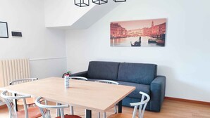 A modern room with a wooden dining table, four chairs, a grey sofa, and a cityscape painting on the wall. - Holiday apartment with balcony (Cavallino-Treporti)