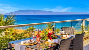 A dining table set with plates, glasses, and a bottle, overlooking a beach and ocean. - Honua Kai | Ocean View 8 BR, Sleeps 26 | Car Incl. w/ 6+ Nights | HKH ML-3095 by KBM (Lahaina)