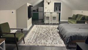 A compact room with a bed, a green armchair, a small desk, a kitchenette, and a TV mounted on the wall. - Moonstruck Studio: In Wine Country (Newberg)
