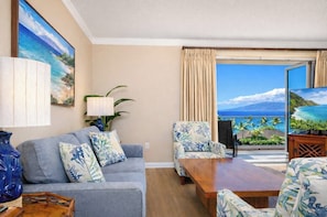 Ein Wohnzimmer mit Sofa, Sesseln, Couchtisch und Fernseher. Ein großes Fenster bietet Blick auf den Ozean.